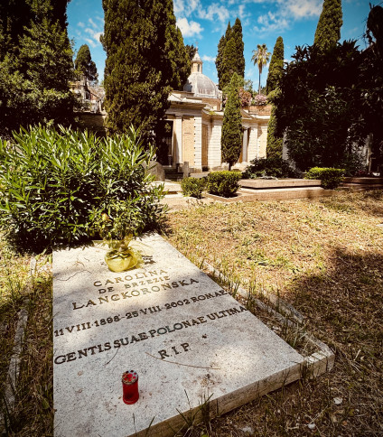 Gravestone of Carolina de Brezie Lanckoronska in a cemetery, surrounded by trees and greenery. The inscription includes her birth and death dates, and a red candle is placed on the grave.