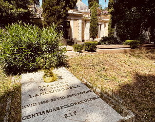 Gravestone of Carolina de Brezie Lanckoronska in a cemetery, surrounded by trees and greenery. The inscription includes her birth and death dates, and a red candle is placed on the grave.