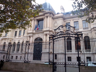 Facade of Hôtel Potocki in Paris with ornate iron gate in the foreground. The building features classical architecture with columns and French flags above the entrance.