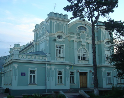 Historyczny budynek z jasnoniebieską fasadą i ozdobnymi białymi detalami, z łukowatymi oknami i dekoracyjnym gzymsem. W pobliżu stoi wysokie drzewo, częściowo zasłaniając widok.