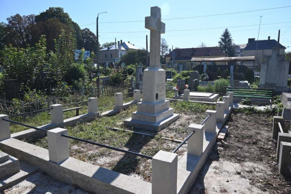 Groby żołnierzy polskich z centralnym kamiennym krzyżem na cmentarzu, otoczone niskimi kamiennymi słupkami i metalowymi prętami. W tle widoczne drzewa i budynki.