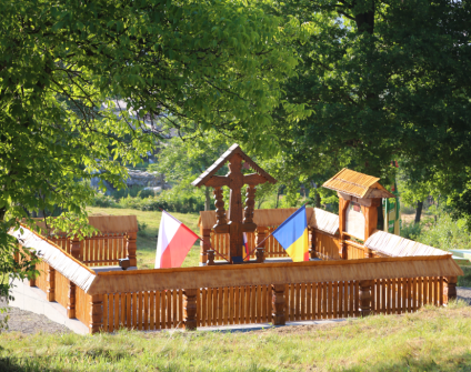 Drewniany pomnik w Berbeşti, Rumunia, z flagami Polski i Rumunii. Konstrukcja w tradycyjnym karpackim stylu, otoczona drewnianym ogrodzeniem i drzewami.