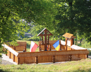 Drewniany pomnik w Berbeşti, Rumunia, z flagami Polski i Rumunii. Konstrukcja w tradycyjnym karpackim stylu, otoczona drewnianym ogrodzeniem i drzewami.