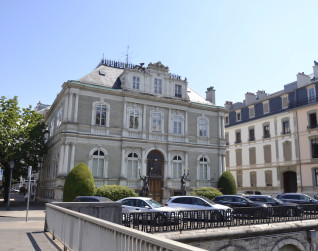 Fotografia budynku Petit Palais w Genewie z kamienną fasadą i dachem mansardowym, ujęcie od ulicy zza metalowego ogrodzenia z zaparkowanymi samochodami przed wejściem