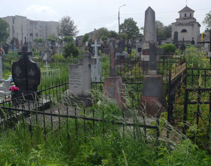 Fotografia grobu z kamiennym krzyzem na cokole i tablica inskrypcyjna, ujetego metalowym ogrodzeniem, wsrod innych nagrobkow