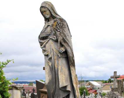 Photograph of a cemetery statue of the Virgin Mary on a rock like pedestal with head bowed right hand on chest left hand holding a lily and an inscription plaque on the front