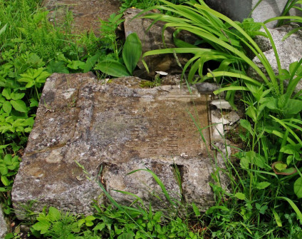 Fotografia przewróconego kamiennego nagrobka wśród trawy i destruktów z prostokątną tablicą w wypukłej ramce i słabo czytelną inskrypcją rytą