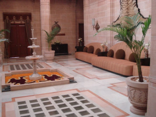 Wnetrze palacu w Dźodhpur w Indiach z dluga tapicerowana sofa i okraglymi poduszkami, palmami w donicach oraz pietrowa szklana fontanna na marmurowej posadzce z geometrycznymi wstawkami
