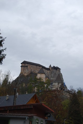 Fotografia Zamku Orawskiego na skalistym wzgórzu ponad drzewami i dachami pod pochmurnym niebem.