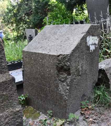 Kamienny cokół z mocno ściętą górną krawędzią, pierwotnie oznaczony krzyżem, obecnie brakującym. Z przodu wyryta inskrypcja. Widoczna otaczająca roślinność i inne nagrobki.