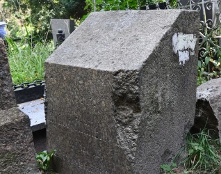 Kamienny cokół z mocno ściętą górną krawędzią, pierwotnie oznaczony krzyżem, obecnie brakującym. Z przodu wyryta inskrypcja. Widoczna otaczająca roślinność i inne nagrobki.
