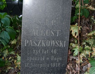 Nagrobek Augusta Paszkowskiego z wyrytą inskrypcją, otoczony zielenią. Kamień ma ścięte górne krawędzie i zwęża się ku górze.