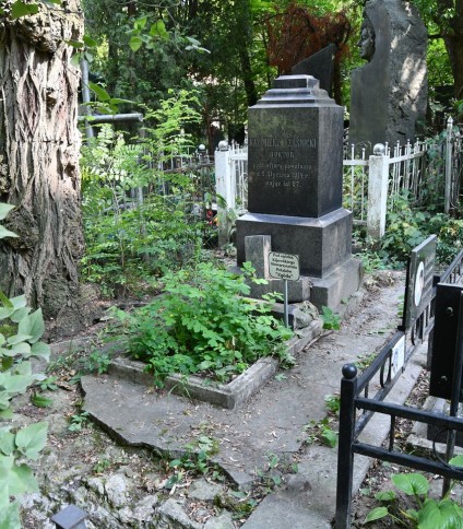 Grób Kazimierza Rząśnickiego z dwustopniowym cokołem i podstawą, z wyrytymi inskrypcjami. Rozczłonkowany krzyż oparty o nagrobek. Otoczony zielenią i pozostałościami ogrodzenia.