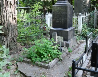 Grób Kazimierza Rząśnickiego z dwustopniowym cokołem i podstawą, z wyrytymi inskrypcjami. Rozczłonkowany krzyż oparty o nagrobek. Otoczony zielenią i pozostałościami ogrodzenia.