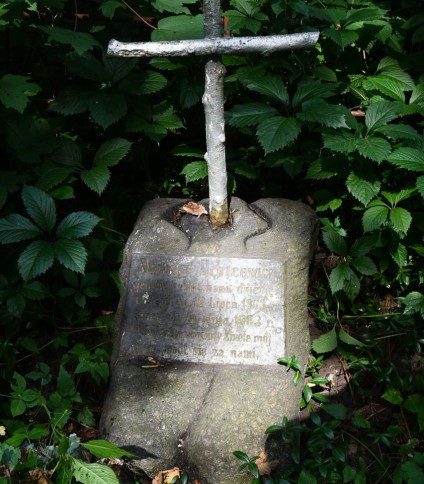 Grób oznaczony krzyżem na cokole. Krzyż przypomina gałęzie drzewa. Cokół to stos kamieni z tablicą z inskrypcją z przodu.