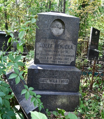 Nagrobek Józefa Pěnički z połamanym krzyżem na trzystopniowym cokole. Inskrypcja w reliefie: 'Józef Pěnička, przeżył 51 lat, zmarł 17 grudnia 1903. Cześć twej pamięci.' Otoczony roślinnością i żelaznym ogrodzeniem.