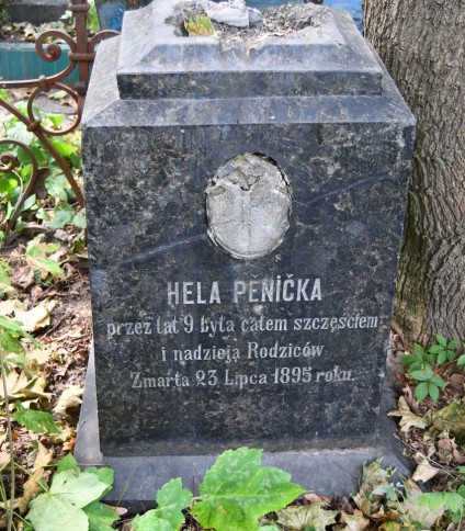 Nagrobek Heli Pěnička z inskrypcją w języku polskim, otoczony roślinnością. Kamień nosi ślady zużycia i ma pozostałości medalionu ze zdjęciem powyżej inskrypcji.