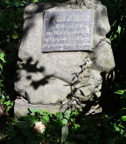 Nagrobek Alfreda Guerquina z krzyżem na kamiennym cokole. Cokół przypomina stos kamieni. Na froncie widoczna tablica z inskrypcją w reliefie.