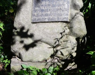 Nagrobek Alfreda Guerquina z krzyżem na kamiennym cokole. Cokół przypomina stos kamieni. Na froncie widoczna tablica z inskrypcją w reliefie.