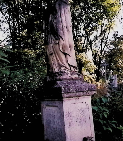 Grób oznaczony figurą Matki Boskiej Różańcowej na trójstopniowym cokole. Matka Boska w geście modlitewnym z różańcem na prawej ręce, ubrana w długą suknię, płaszcz i welon.