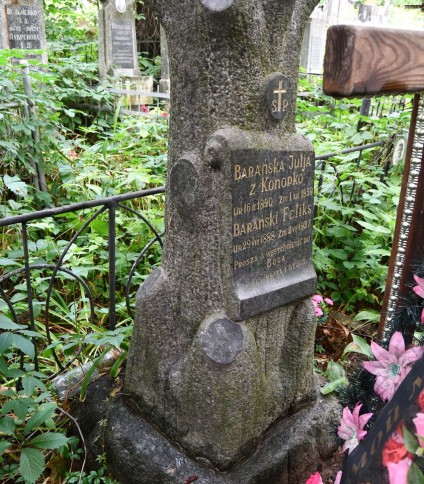 Nagrobek Feliksa i Julii Barańskich z cokołem w kształcie pnia drzewa i grawerowanymi tablicami. Otoczony metalowym ogrodzeniem ze szpiczastymi słupkami. Kwiaty i zieleń wokół.