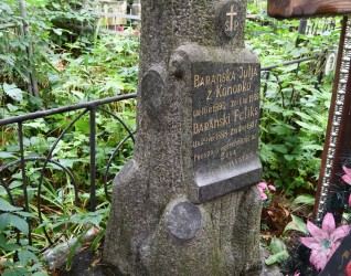 Nagrobek Feliksa i Julii Barańskich z cokołem w kształcie pnia drzewa i grawerowanymi tablicami. Otoczony metalowym ogrodzeniem ze szpiczastymi słupkami. Kwiaty i zieleń wokół.