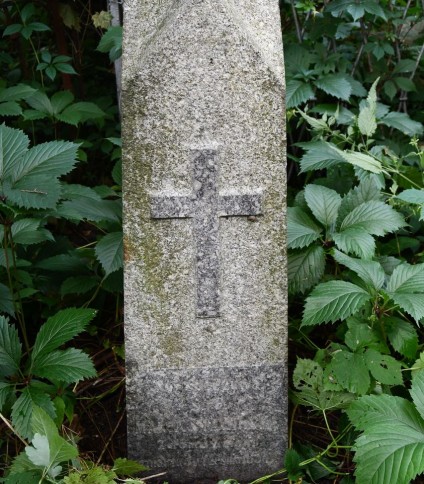 Kamienny cokół z reliefowym krzyżem i inskrypcją, otoczony zielenią. Cokół zwęża się ku górze i jest zwieńczony daszkowatą strukturą.