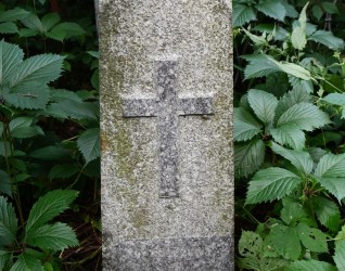 Kamienny cokół z reliefowym krzyżem i inskrypcją, otoczony zielenią. Cokół zwęża się ku górze i jest zwieńczony daszkowatą strukturą.