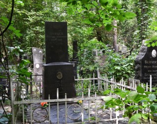 Grób Stanisława Pawłowskiego z krzyżem na cokole, otoczony metalowym ogrodzeniem z okrągłymi wzorami. Teren porośnięty zielenią i drzewami.