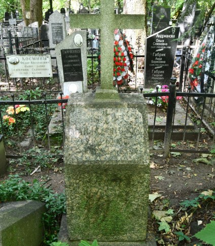 Nagrobek Marii Smulskiej z krzyżem na cokole, z zwężającym się cokolikiem i inskrypcją w reliefie z ściętymi narożnikami. Otoczony innymi nagrobkami i zielenią.