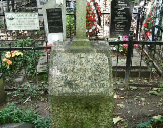 Nagrobek Marii Smulskiej z krzyżem na cokole, z zwężającym się cokolikiem i inskrypcją w reliefie z ściętymi narożnikami. Otoczony innymi nagrobkami i zielenią.