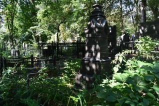 Nagrobek Marcina Gaczkowskiego z krzyżem na stopniowanej podstawie, otoczony metalowym ogrodzeniem z zakończonymi grotami słupkami. Teren jest bujny w zieleń, w tle widoczne inne nagrobki.