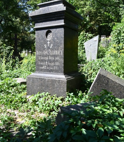 Nagrobek Bazylego Ustyanowicza z obeliskiem leżącym obok. Podstawa ma ścięte krawędzie i inskrypcję z przodu. Teren porośnięty zielenią.