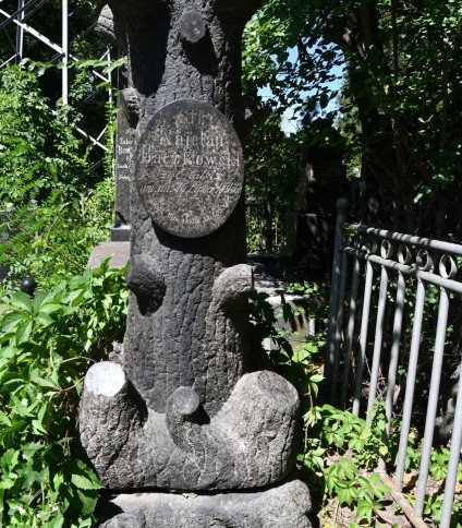 Nagrobek Kajetana Baczkowskiego z podstawą stylizowaną na korzenie drzewa i cokołem jako pień drzewa. Z przodu owalna tabliczka z rytą inskrypcją. Otoczony zielenią i metalowym ogrodzeniem.