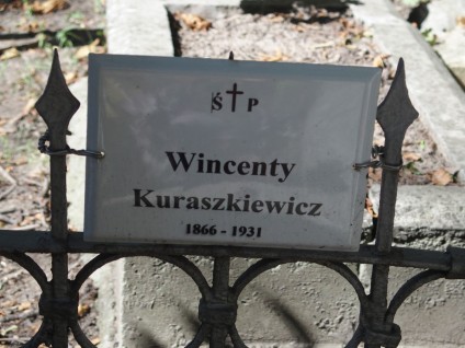 Metalowa tabliczka na ogrodzeniu grobu z napisem 'Wincenty Kuraszkiewicz 1866-1931'. Ogrodzenie ma ostro zakończone pręty i okrągłe wzory.