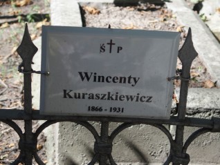 Metalowa tabliczka na ogrodzeniu grobu z napisem 'Wincenty Kuraszkiewicz 1866-1931'. Ogrodzenie ma ostro zakończone pręty i okrągłe wzory.