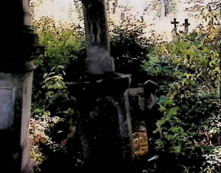 Nagrobek Teodora Skrockiego z krzyżem na dwustopniowym cokole, z reliefem Chrystusa na przecięciu ramion. Otoczony roślinnością.