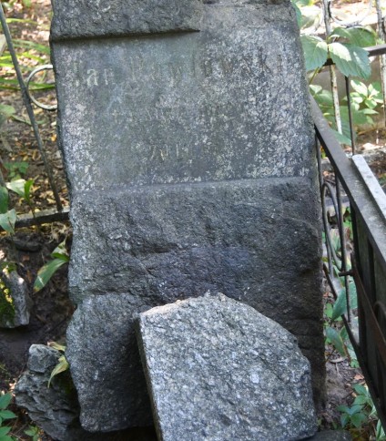 Kamienny cokół z inskrypcją dla Jana Pawłowskiego, bez krzyża. Cokół to nieobrobiony blok z wyrytym tekstem z przodu. Otoczony roślinnością i metalowym ogrodzeniem.