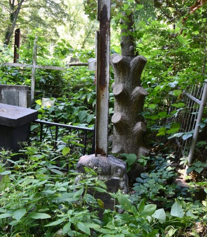 Grób Marii i Mieczysława Korzeniowskich z krzyżem stalowym na cokole przypominającym stos kamieni. Krzyż bez poprzecznej belki. Otoczony gęstą zielenią.