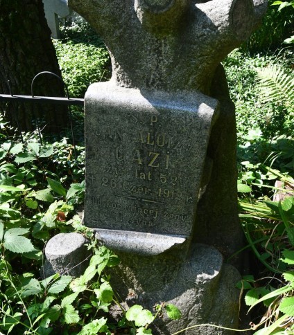 Nagrobek Jana-Aloizego Gazi z cokołem w kształcie pnia drzewa i tablicą reliefową z jego imieniem i datami. Otoczony zielenią.
