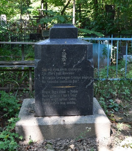 Nagrobek z inskrypcją na cokole i podstawie, otoczony zielenią. Cokół ma fazowane krawędzie, w tle widoczny krzyż.