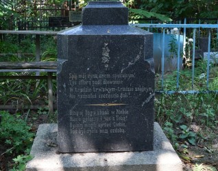 Nagrobek z inskrypcją na cokole i podstawie, otoczony zielenią. Cokół ma fazowane krawędzie, w tle widoczny krzyż.