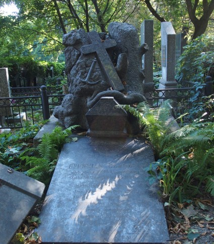 Grób Cypriana Ułaszyna z ukośną płytą nagrobną i krzyżem na dwustopniowym cokole. Cokół przypomina stos kamieni z pniem drzewa. Widoczny relief krzyża i kotwicy, trumna z wężem oraz paprocie.