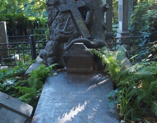 Grób Cypriana Ułaszyna z ukośną płytą nagrobną i krzyżem na dwustopniowym cokole. Cokół przypomina stos kamieni z pniem drzewa. Widoczny relief krzyża i kotwicy, trumna z wężem oraz paprocie.