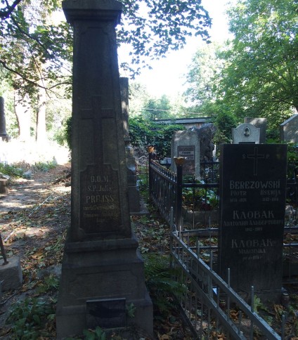 Nagrobek Julii Prejss z dwustopniowym cokołem i podstawą, pierwotnie oznaczony krzyżem. Z przodu tablica z inskrypcją w formie krzyża. Widoczne otaczające groby i drzewa.