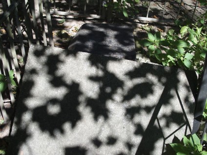 Nagrobek Katarzyny Stanisławskiej z cokołem o ściętych górnych krawędziach i podstawie. Cokół leży przewrócony obok podstawy, otoczony zielenią i cieniami.