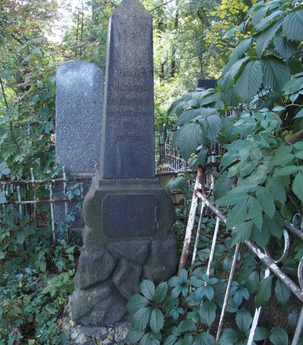 Nagrobek Olimpii i Konrada Żendzianowskich z obeliskiem na kamiennej podstawie, otoczony zielenią i żelaznym ogrodzeniem. Widoczna inskrypcja z przodu.