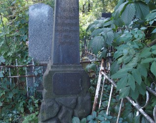 Nagrobek Olimpii i Konrada Żendzianowskich z obeliskiem na kamiennej podstawie, otoczony zielenią i żelaznym ogrodzeniem. Widoczna inskrypcja z przodu.