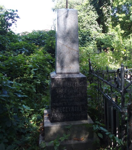 Nagrobek Eweliny i Adama Męczyńskich. Brak oryginalnego krzyża, widoczny żeliwny trzpień. Cokół dwustopniowy, pierwszy stopień o ściętych krawędziach, drugi zwężający się ku górze. Otoczony zielenią.