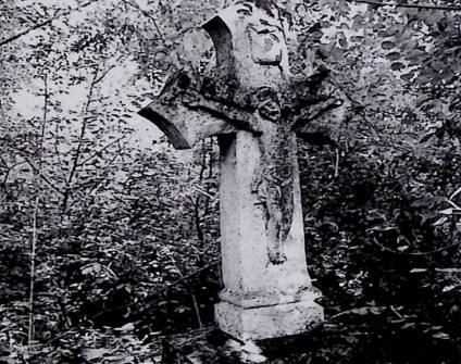 Nagrobek z krzyżem na dwustopniowym cokole otoczony roślinnością. Krzyż z ramionami zakończonymi wykrojem łuku kotarowego, z reliefem Chrystusa na przecięciu i titulus powyżej.